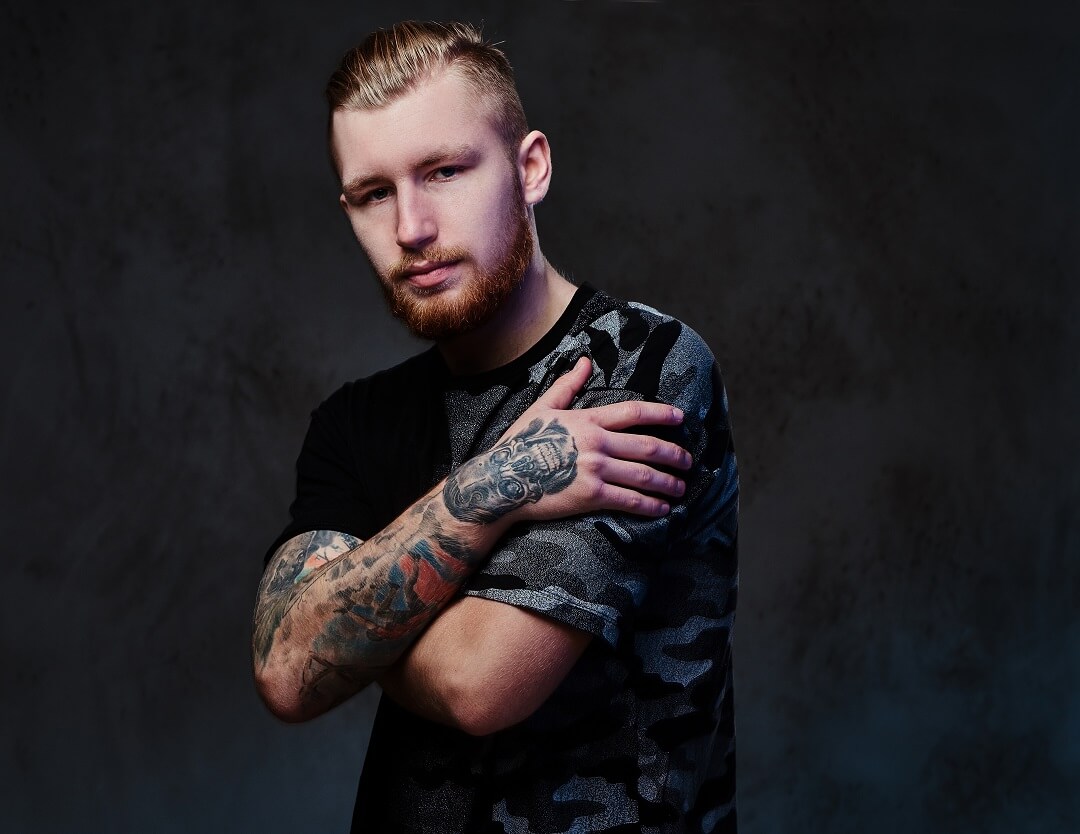 Redhead bearded young male with tattoos on arms, dressed in a black t shirt over dark grey background.